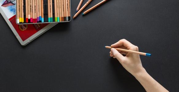 A hand holding a blue coloured pencil poised above a black sheet of paper.