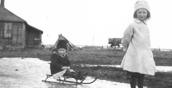 Black and white photograph of a young girl pulling a child on a wooden toboggan. 