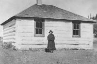 Black and white photo of a woman standing in front of a one-room schoolhouse. 