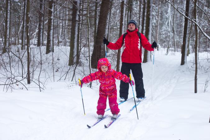 young girl learning to cross-country ski with her dad through trees