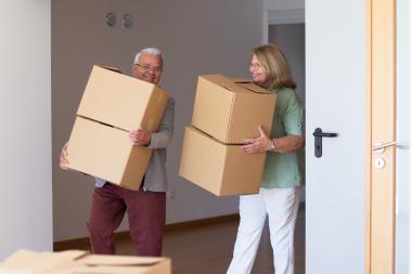 A male senior and female senior both smile as they carry brown moving boxes 